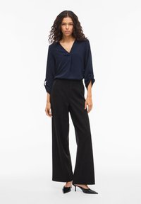 Μπλε μπλούζα με μακριά μανίκια, V-λαιμόκοψη και γυρισμένα μανίκια, συνδυασμένη με μαύρο παντελόνι φαρδιών ποδιών και μυτερά μαύρα ψηλοτάκουνα. Απλός, δομημένος σχεδιασμός.