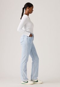 Γυναίκα με λευκό μακρυμάνικο πουκάμισο, ανοιχτού μπλε τζιν και λευκά αθλητικά παπούτσια με πράσινο σήμα, στέκεται προφίλ με τα χέρια στις τσέπες.