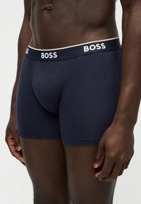 Homme portant un boxer bleu marine avec le logo "BOSS" sur la ceinture élastique, debout devant un fond clair uni.