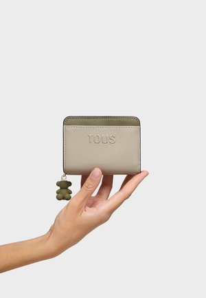 Mano che tiene un piccolo portafoglio beige e oliva con il marchio "TOUS" impresso e un ciondolo a forma di orsetto oliva come tiralampo, su sfondo bianco.