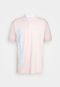 Polo rose pâle à manches courtes avec une patte à deux boutons, logo "Lacoste" vertical bleu clair sur le côté gauche, et petit emblème de crocodile sur la poitrine.