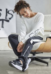 White sweatshirt with "CO" on the sleeve, black joggers with stripes and text, black sneakers, seated in a mid-century style chair.