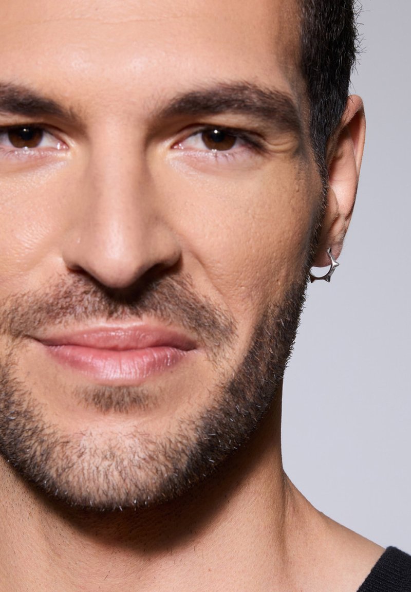 Srebrna, gwiazdkowa kolczyk na lewym uchu, gładka skóra, subtelne owłosienie na twarzy i neutralne tło.