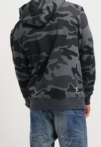 Mikina s kapucí v černém a šedém camo vzoru, vyrobená z jemné tkaniny, s žebrovanými manžetami a šňůrkou na kapuci.