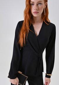 Blusa nera avvolgente realizzata in tessuto morbido, con maniche lunghe, scollatura a V e dettaglio a nodo in vita, abbinata a una pochette.