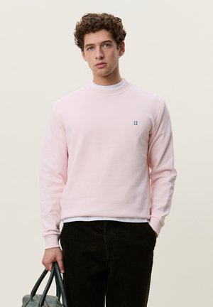 Jeune homme aux cheveux bouclés portant un sweat-shirt rose clair et un pantalon noir, tenant un sac à main vert foncé dans une main.