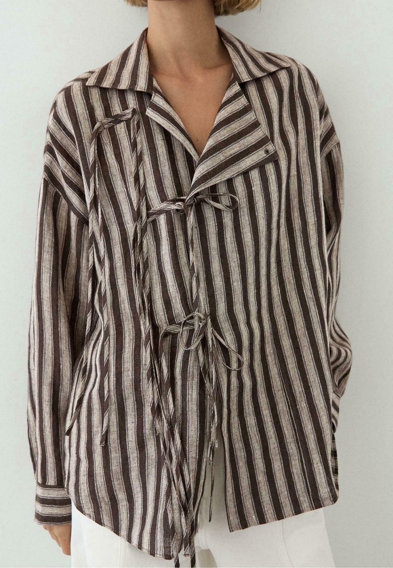 Personne portant une veste oversize à rayures verticales marron et beige avec fermetures à lacets et un pantalon blanc sur un fond uni.