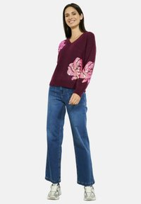 Maglione bordeaux scuro a scollo a V con grandi paillettes a forma di fiore rosa sulle maniche, indossato con jeans a gamba larga blu chiaro e sneakers.