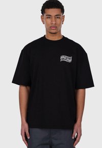 Zwart T-shirt met korte mouwen van katoen. Heeft een witte bedrukte logo op de linkerkant van de borst. Ontspannen pasvorm met een ronde hals.