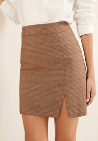 Femme portant une mini-jupe à carreaux bruns taille haute avec une petite fente devant, assortie à un chemisier blanc à manches longues.