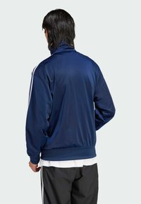 Veste de survêtement bleu marine en tissu lisse, dotée d'un col montant, de bandes latérales blanches et de poignets élastiques avec un détail au bas.