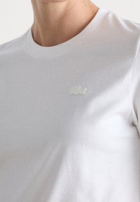 White cotton t-shirt with a crew neckline and a small, embroidered light-colored Lacoste logo on the chest. Smooth texture.