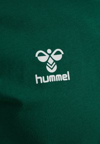 Grönt tyg med en slät struktur pryds av en framträdande vit logotyp med stiliserade inslag och ordet "hummel" under.
