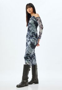 Lang off-shoulder jurk in een blauw en grijs bloemenprint, aangesloten figuur, met lange mouwen. Gecombineerd met kniehoge zwarte leren laarzen.