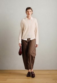 Beige strikkegenser over en hvit skjorte med krage, brune vide bukser og maroon loafers, holder en liten maroon håndveske.