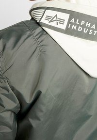 Oliivinvihreä pommikonejacketti, jossa on kiiltävä pinta, valkoisella fleece-vuorella varustettu huppu ja raidallinen painettu kauluslabeli, jossa on logo.