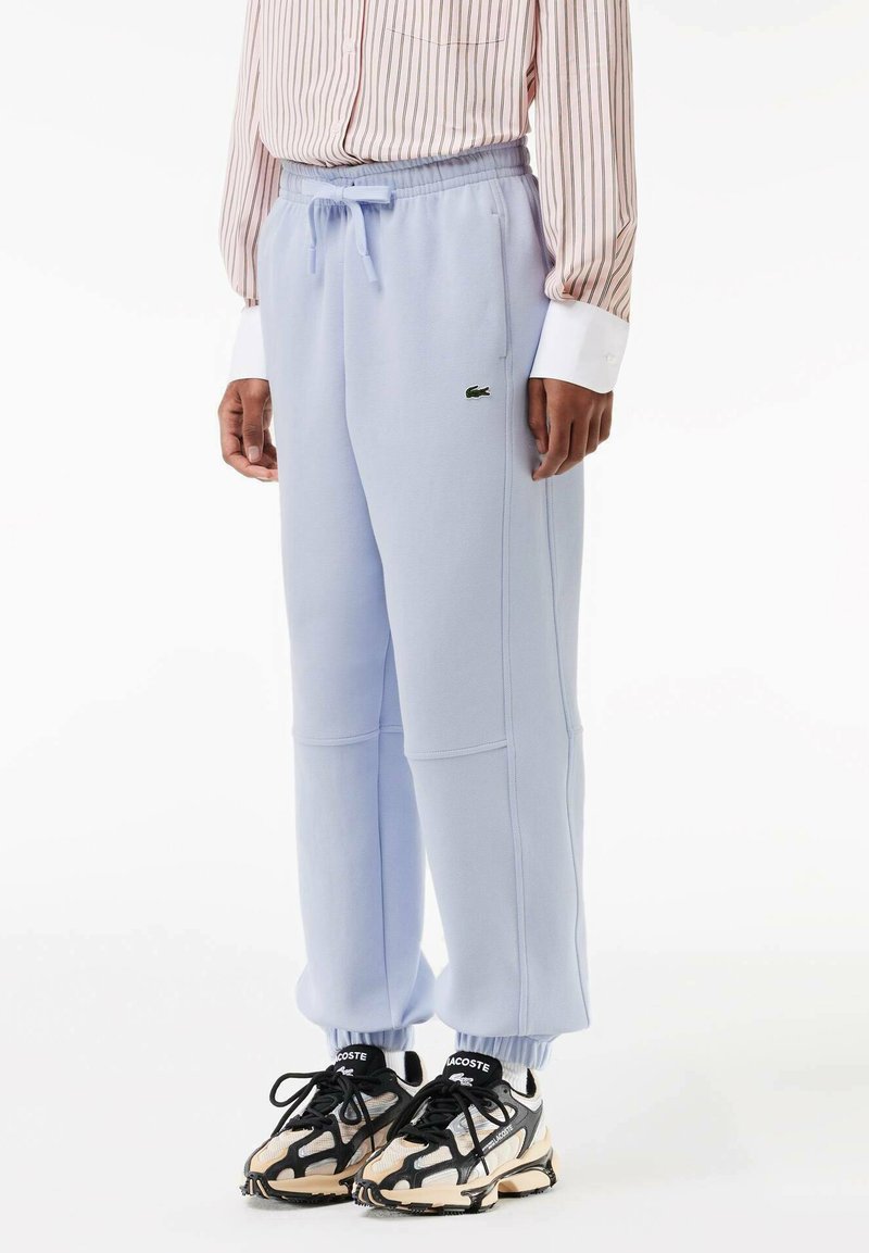 Pantalones de chándal azul claro con cinturilla y puños elásticos, que presentan un pequeño logo verde de Lacoste en el lateral. Se combinan con zapatillas negras y crema.