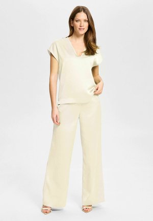 Mujer de pie con pantalones anchos color crema, camiseta de manga corta con cuello en V y tacones abiertos dorados sobre un fondo claro y liso.
