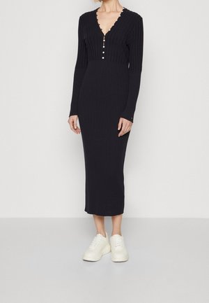 Vrouw die een nauwsluitende zwarte ribgebreide midi-jurk draagt met lange mouwen, een V-hals met kleine knoopjes en witte sneakers, staand tegen een effen achtergrond.