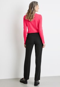 Figurbetonter, pinkfarbener Pullover mit langen Ärmeln, der über Knopfdetails an den Manschetten verfügt. Kombiniert mit schwarzen, maßgeschneiderten Hosen und schwarzen Schuhen.