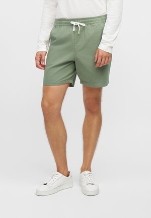 Un mannequin porte un short vert sauge à cordon de serrage, une chemise blanche à manches longues et des baskets blanches sur un fond uni.