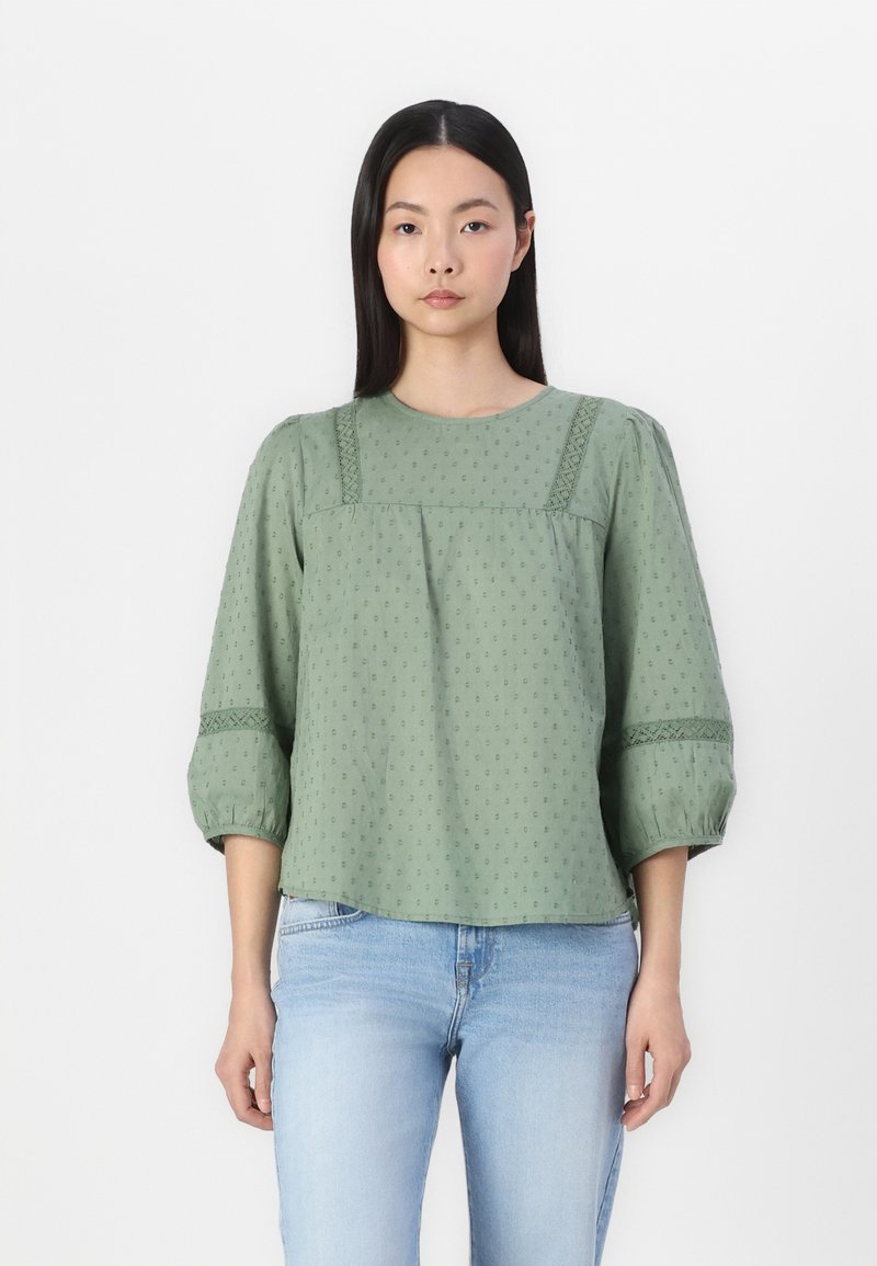 Blusa verde realizzata in tessuto strutturato con scollatura rotonda, maniche a sbuffo e dettagli ricamati lungo il petto e le maniche.
