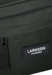 Sötétzöld szövet táska fekete cipzárral és fekete címkével, fehér felirattal: "LARKSON HAMBURG". Simább textúrával és tartós kivitelezéssel rendelkezik.
