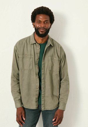 Olive green button-up shirt, two chest pockets, lightweight cotton fabric, relaxed fit, paired with a dark green T-shirt underneath.