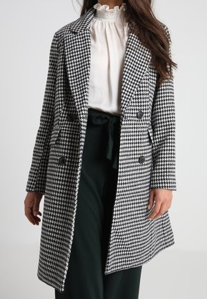 Zwart-wit houndstooth-gepatenteerde jas met een dubbelzijdig ontwerp, brede revers en twee voorzakken. Voorzien van grote zwarte knopen.