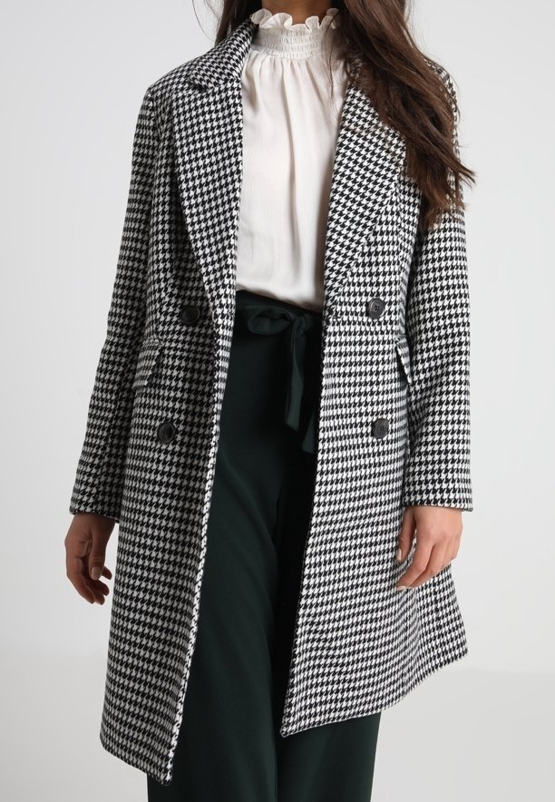 Schwarz-weißer Mantel im Pepita-Muster mit doppelter Brust, breiten Revers und zwei Fronttaschen. Ausgestattet mit großen schwarzen Knöpfen.