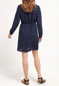 Vestido azul marino con mangas largas, cintura ceñida con cinturón y textura suave, combinado con sandalias negras que tienen suela de corcho y tiras.