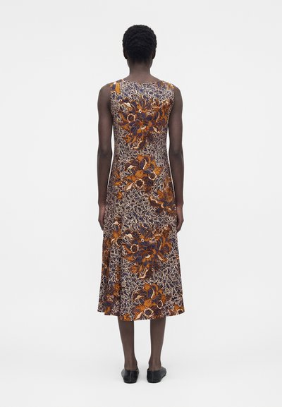 Γυναίκα που στέκεται με την πλάτη, φοράει μίντι φόρεμα χωρίς μανίκια με πορτοκαλί, μπλε και λευκό floral σχέδιο, συνδυασμένο με μαύρα παπούτσια χωρίς κορδόνια.