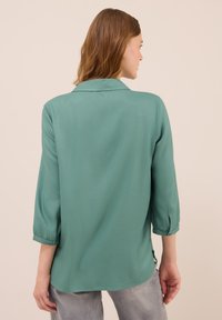 Frau mit hellbraunem Haar, die eine türkisfarbene Bluse mit langen Ärmeln und eine hellgraue Jeans trägt, von hinten betrachtet vor einem einfarbigen hellbeigen Hintergrund.