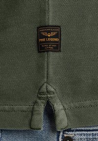 Olivově zelené úpletové tričko s texturovaným vzhledem, které má černou nášivku s označením "PME Legend" a unikátní detail bočního rozparku.