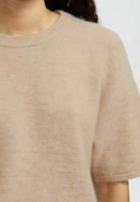 Pull en tricot beige à manches courtes, col rond et texture douce. Le tissu semble léger et légèrement duveteux avec un éclat subtil.