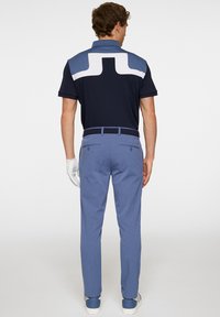 Marineblaues Poloshirt mit einem weißen und hellblauen geometrischen Design auf dem Rücken, kombiniert mit hellblauen Hosen, getragen mit einem Handschuh und Turnschuhen.