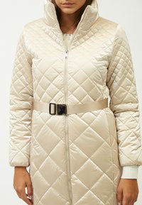 Abrigo largo acolchado en beige claro, fabricado en un material brillante. Cuenta con un cuello alto, cremallera frontal y un cinturón a juego en la cintura.