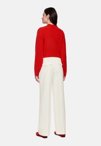 Roter kurzer Pullover mit gerippter Struktur, kombiniert mit hoch taillierter weißer Hose mit Taschen. Modell steht mit dem Rücken zur Kamera.