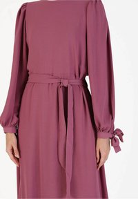 Robe rose à manches longues en tissu doux avec une ceinture à la taille et des accents de nœud sur les manches. Texture lisse et design simple.