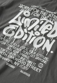 Graues Baumwoll-T-Shirt mit weißen grafischen Texten, das fettgedruckte, stilisierte Typografie und verschiedene Botschaften zeigt. Weicher Stoff mit Siebdruckdetails.