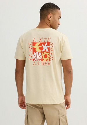 Hombre con camiseta beige con un gráfico rojo y amarillo de pájaros, conchas marinas, estrella de mar y sol con texto en francés en la parte trasera.