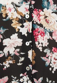 Svart blus med ett blommönster i nyanser av rosa, kräm och blått. Pärlknappar pryder framsidan och ger en mjuk textur.