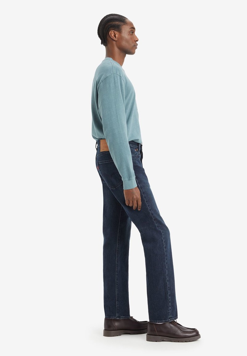 Turquoise longhulshemd met ribbelmanchetten, gecombineerd met donkerblauwe rechte jeans met een contrasterend bruin leren patch aan de achterkant.