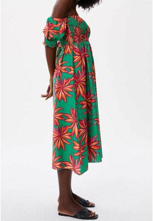Vrouw draagt een groene midi-jurk met oranje en roze bloemenpatroon en off-shoulder pofmouwen, gecombineerd met zwarte slides.
