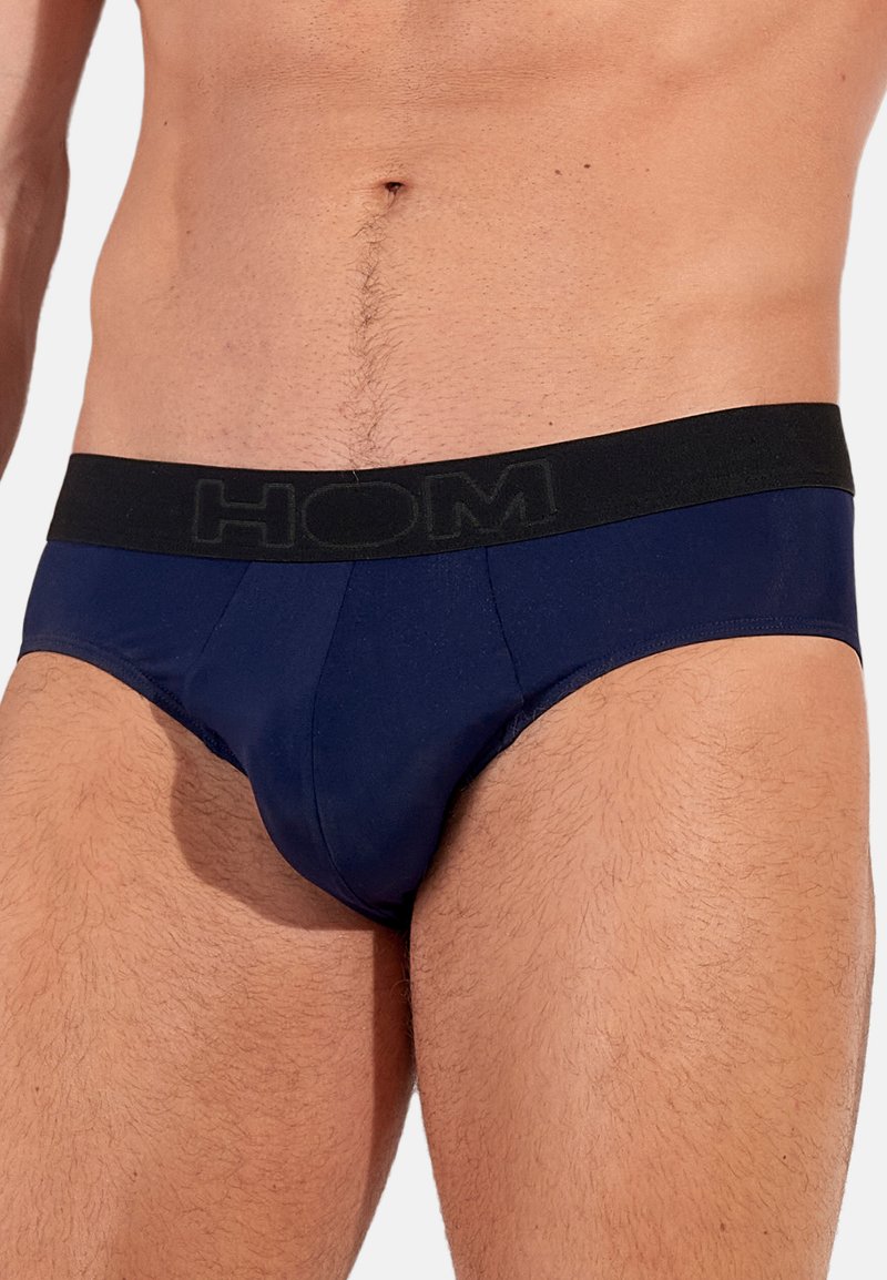 Marineblauwe slips met een zwarte elastische tailleband waarop het merk "HOM" in grijs is gedrukt. Glad materiaal, strakke pasvorm en minimale stikseldetails.