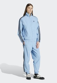 Lichtblauwe trainingspak met een ritsjack en broeken met een elastische taille, afgewerkt met zwarte strepen en een geborduurd logo op de borst.