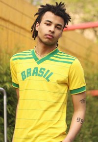 Gul t-shirt med gröna ränder och texten "BRASIL". Kort ärm, v-ringning, gjort av mjukt tyg. Visar en tatuering på armen.