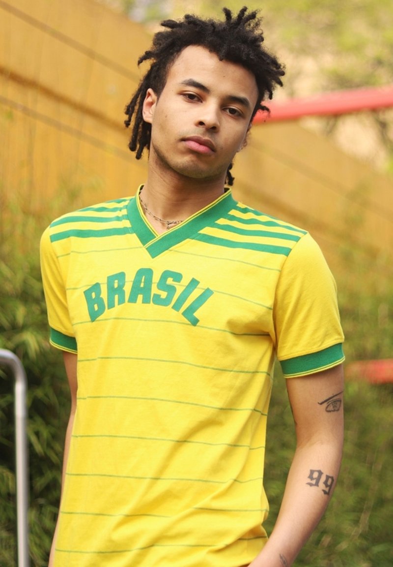 Gul t-shirt med gröna ränder och texten "BRASIL". Kort ärm, v-ringning, gjort av mjukt tyg. Visar en tatuering på armen.