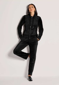 Vrouw in een zwart velours trainingspak met een subtiele jacket met luipaardprint, staande op één been tegen een eenvoudige lichte achtergrond.