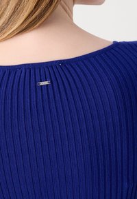 Gros plan sur le haut du dos d'une personne portant un haut en maille côtelée bleue avec une petite étiquette de marque argentée rectangulaire près de l'encolure.